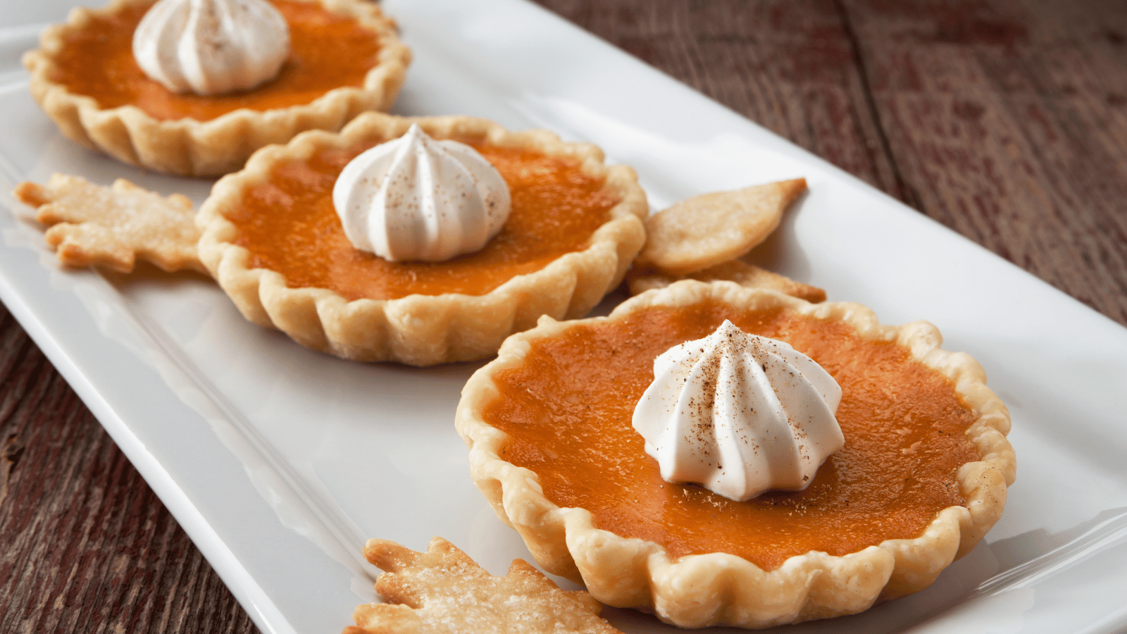 Three pumpkin tarts with dollop of cream on top on top of white platter with leaf dough cutouts
